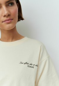 Hanorac din bumbac de culoare crem, supradimensionat, cu mâneci scurte, având brodat în negru textul "Sans effort, chic et class" și "FEMME."