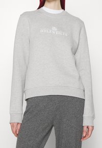 Svetle šedá mikina s žebrovanými manžetami a lemem. Má bílé logo vytištěné na hrudi. Měkká textura, volný střih, minimalistický design.