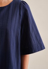 Top blu navy con maniche corte a sbuffo, decorato con ricami verticali e scollatura rotonda, realizzato in tessuto leggero con una finitura testurizzata.