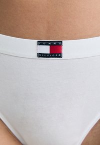 Bikini en coton blanc avec une texture lisse et une taille de marque arborant un logo rectangulaire dans des couleurs bleu marine, rouge et blanc.