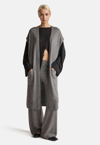 Šedý pletený kardigan s otevřeným předním dílem a kapsami, spárovaný s odpovídajícími volnými šedými kalhotami. Černý top s dlouhým rukávem pod tím. Hladká textura.