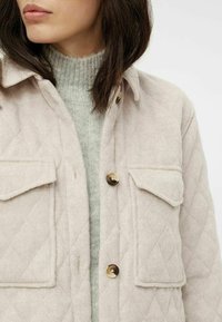 Chaqueta acolchada de color rosa claro con dos bolsillos en el pecho y botones dorados, superpuesta sobre un suéter de canalé gris claro. Textura suave.