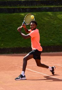 Tennisspiller i rosa skjorte og svarte shorts svinger en racket mot en gul ball. Banen er rød leire med grønt gress i bakgrunnen.