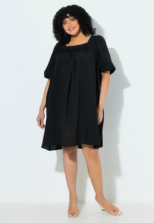 Donna con capelli ricci che indossa un vestito nero largo fino al ginocchio con maniche a sbuffo e tacchi beige con punta aperta, in piedi su uno sfondo bianco.