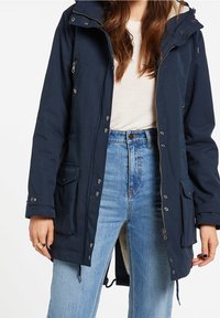 Mujer vestida con una chaqueta con capucha de color azul marino y grandes bolsillos, sobre una camiseta blanca y vaqueros de cintura alta de color azul claro.