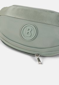 Marsupio in nylon verde con una tracolla testurizzata a righe, logo circolare con "B" e chiusura laterale con zip. Superficie liscia e forma arrotondata.