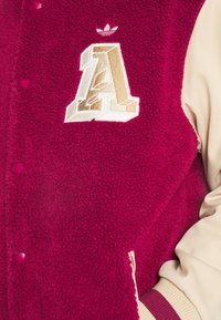 Vinröd fleecejacka med beigea ärmar, med en framträdande guldfärgad och vit broderad bokstav "A" och en Adidas-logotyp.