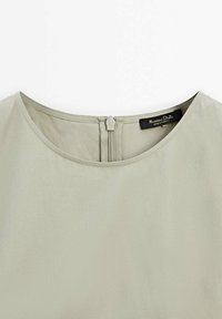 Haut beige clair avec un col rond et une fermeture éclair centrée au dos, montrant l'étiquette de la marque cousue à l'intérieur du col.