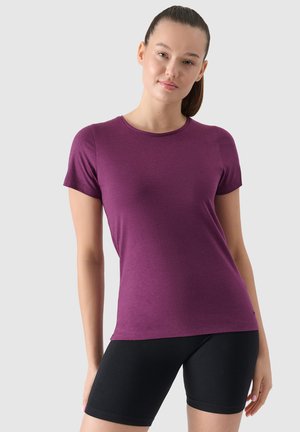 Mujer con el cabello recogido que lleva una camiseta ajustada de manga corta color ciruela y pantalones cortos de ciclismo negros sobre un fondo claro y liso.