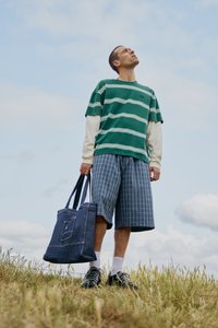 Striped green and white short-sleeve shirt over a long-sleeve white shirt, plaid shorts, denim tote bag, and sneakers on grass.