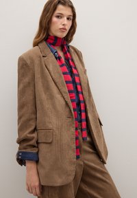 Bruine corduroy blazer met brede revers. Gelaagd met een blauwe shirt en een rood-blauwe gestreepte sjaal. Glad textuur, nauwsluitende pasvorm.