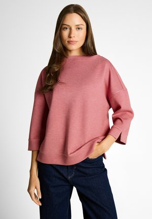 Jonge vrouw met lang bruin haar, gekleed in een losse roze top met lange mouwen en donkerblauwe spijkerbroek, hand in de zak, neutrale achtergrond.