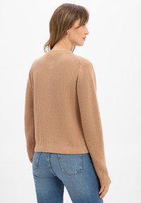 Frau trägt einen beigen, gerippten Strickpullover und blaue Jeans, seitlich gedreht, vor einem schlichten weißen Hintergrund.