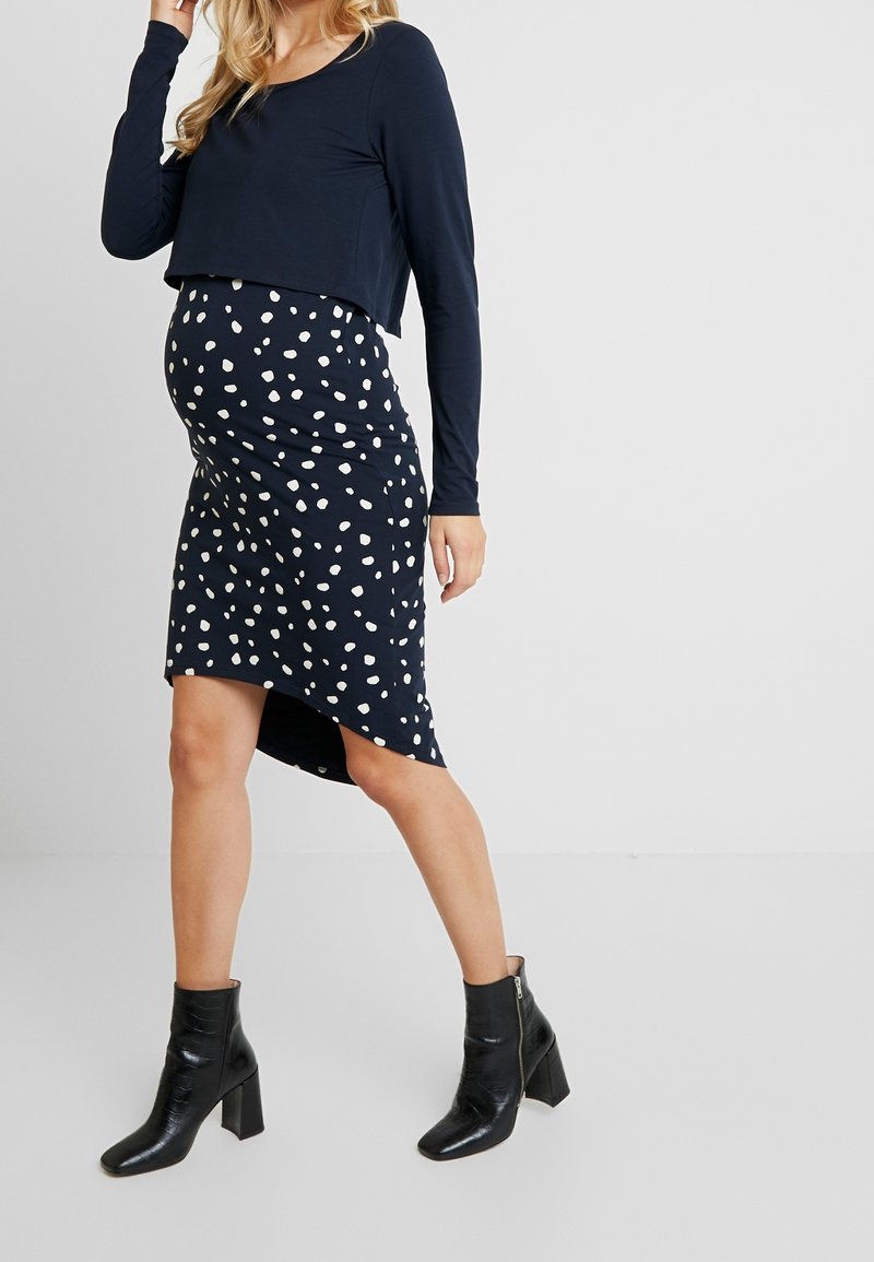 Femme enceinte vêtue d'un haut bleu marine à manches longues, d'une jupe bleu marine à pois blancs et de bottines noires, debout sur un fond uni.