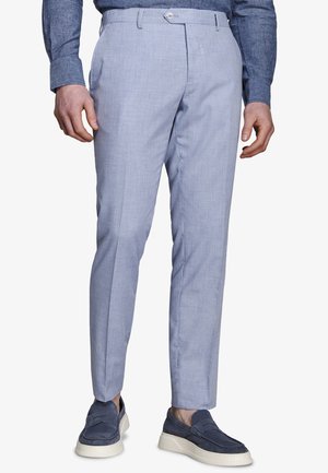 Mężczyzna ubrany w jasnoniebieskie obcisłe spodnie, ciemnoniebieską koszulę z długim rękawem oraz granatowe zamszowe mokasyny z białymi podeszwami, stojący.