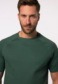 Camiseta de manga corta, color verde oscuro, fabricada en un material suave. Presenta un clásico cuello redondo y mangas raglán con una textura lisa.