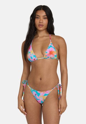 Donna che indossa un bikini tie-dye floreale colorato con top a fascia e slip con laccetti laterali, in piedi davanti a uno sfondo chiaro e semplice.