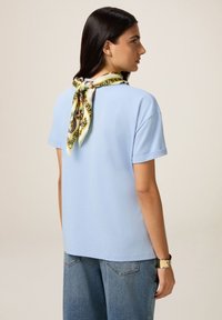 Camiseta de algodón azul claro con mangas cortas, que presenta un diseño de mangas enrolladas y una bufanda amarilla y verde con patrón atada al cuello.
