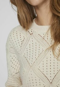 Sweter dzianinowy w kolorze kremowym z geometrycznymi wzorami ażurowymi, prążkowanym dekoltem i zróżnicowaną teksturą materiału.