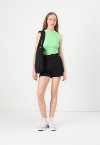Grønn ribbet ermeløs crop topp, svarte belte shortser og hvite høy-top sneakers. Modellen holder en svart skulderveske. Enkel bakgrunn.
