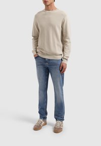 Beige gebreide trui met een gestructureerd patroon, gecombineerd met blauwe jeans en bruine sneakers met witte strepen. Casual fit en ontspannen stijl.
