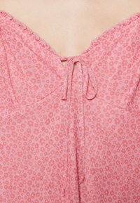 Růžová halenka s květinovým vzorem, kterou zdobí uvázaný výstřih, volánkové okraje a lehký materiál, navržená pro pohodlný střih.