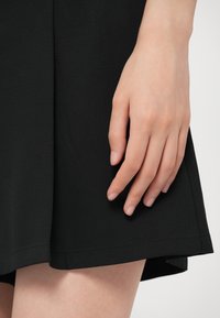 Hand rustend naast de zoom van een zwarte rok, met een gladde stoftextuur en een natuurlijke huidtint op een lichte achtergrond.