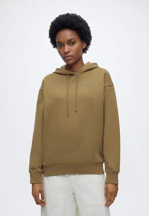 Vrouw met kort krullend haar die een olijfgroene hoodie en witte broek draagt, staand tegen een effen witte achtergrond.