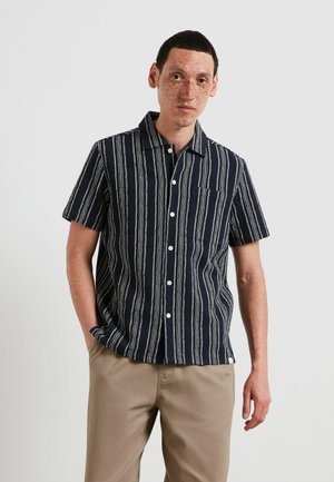 Uomo con lentiggini che indossa una camicia a maniche corte a righe blu navy e bianche con bottoni e pantaloni beige, in piedi davanti a uno sfondo semplice.