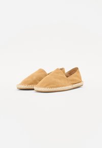 Chaussures slip-on en suede beige avec un bout arrondi et une semelle tressée de type espadrille. Surface texturée, design minimaliste et sans matériel visible.