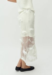 Falda blanca transparente con bordado floral, textura similar a la organza, ligeramente más larga en la parte trasera, combinada con zapatos planos negros.