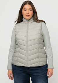 Jasnoszara kamizelka puchowa z wysokim kołnierzem, wzorem w romby, zamek błyskawiczny oraz boczne kieszenie, noszona na jasnoszarym longsleeve.