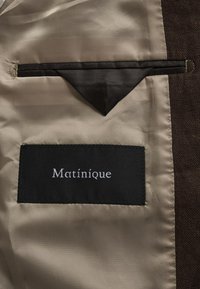 Étiquette noire "Matinique" cousue à l'intérieur de la doublure satinée beige d'une veste marron, avec un rabat de poche triangulaire au-dessus de l'étiquette.