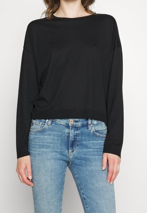 Frau trägt ein schwarzes langärmeliges Oberteil und hellblaue Jeans, steht vor einem einfarbigen weißen Hintergrund.