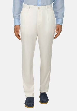 Homme portant un pantalon blanc sur mesure, une chemise bleue boutonnée et des mocassins bleu foncé, debout devant un fond uni.