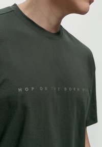 Temno zeleni bombažni T-majica z okroglim izrezom, kratkimi rokavi in tiskanim besedilom "HOP ON THE BORN WAY" na sprednji strani.