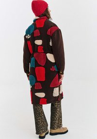 Osoba oblečená v farebnom fleece kabáte z patchworku, červenej čiapke, nohaviciach s leopardím vzorom a béžových turistických topánkach, otočená chrbtom na jednoduchom pozadí.