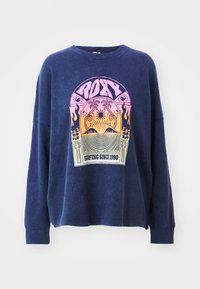 Sweat-shirt surdimensionné bleu marine en tissu texturé, avec un imprimé graphique coloré comportant le texte "ROXY" et des éléments décoratifs.