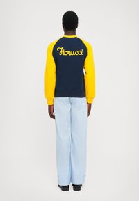 Person kledd i en marineblå og gul sweatshirt med "Fiorucci" på ryggen, lyseblå stripete bukser, stående med ryggen til på en ensfarget bakgrunn.