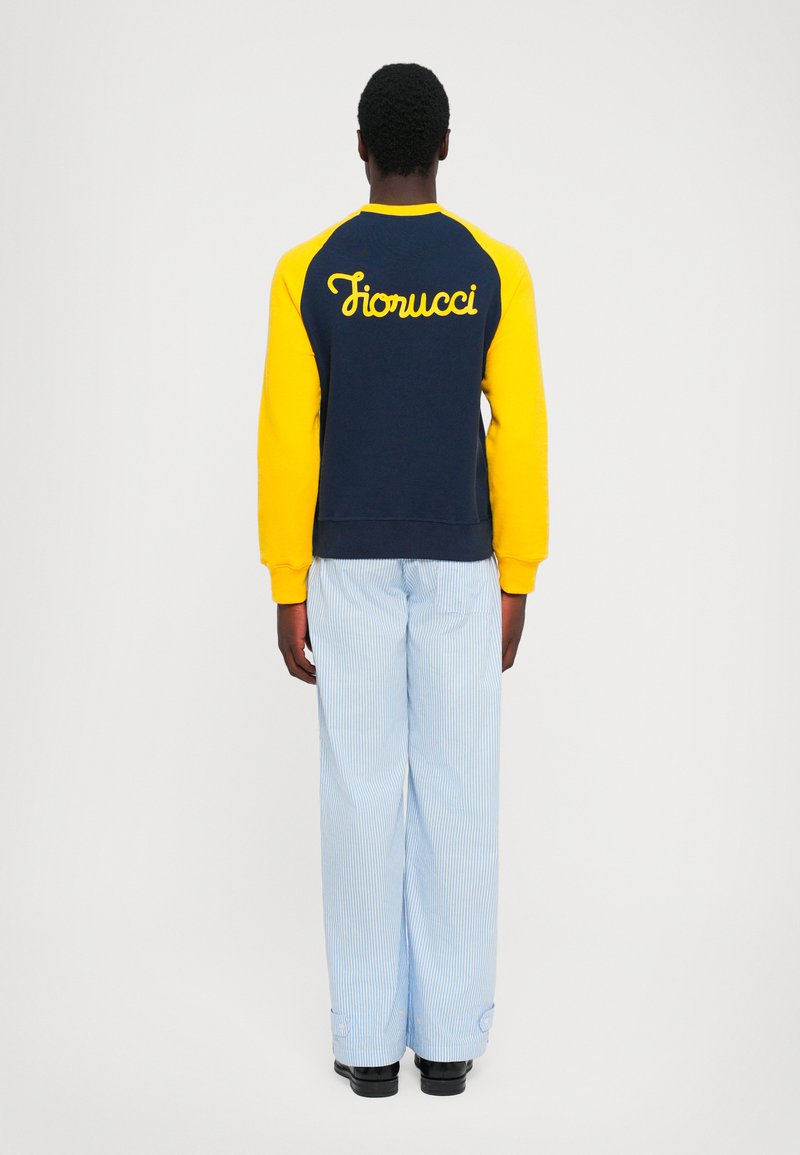 Person kledd i en marineblå og gul sweatshirt med "Fiorucci" på ryggen, lyseblå stripete bukser, stående med ryggen til på en ensfarget bakgrunn.