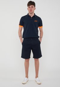 Polo shirt navy con accenti arancioni, con colletto e maniche corte, abbinato a pantaloncini navy. Il modello è in piedi con le mani nelle tasche.