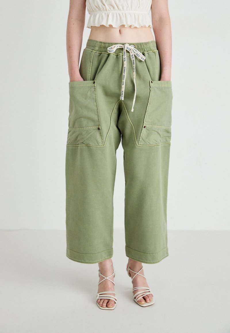 Person i bredde bukser i olivengrøn med store frontlommer, en hvid cropped top og strappy hvid høje hæle sandaler.