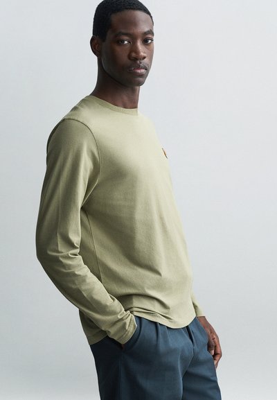 Long-sleeve olive green shirt made from soft cotton, featuring a round neckline and a small chest logo, paired with dark blue trousers.