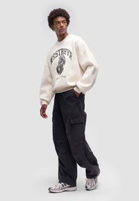 Felpa crema con testo grafico e disegno, abbinata a pantaloni cargo neri larghi e scarpe sportive argento. Collo rotondo e vestibilità oversize.