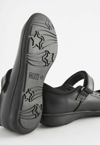 Zapatos de cuero negro con una correa de velcro, que presentan una suela de goma texturizada con patrones de estrellas grabados. Diseño sencillo y redondeado.