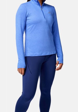 Vrouw draagt een lichtblauw lang mouwtje met rits en duimgaten, samen met donkerblauwe aansluitende legging, staand tegen een effen achtergrond.