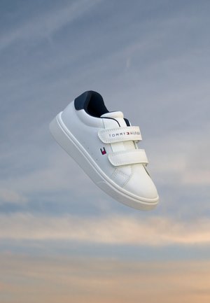 Basket blanche avec double bride velcro et doublure intérieure noire, flottant sur un fond de ciel bleu et orange doux.