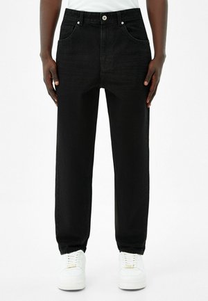 Personne portant un jean droit noir et des baskets blanches, debout devant un fond blanc uni.