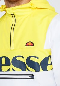Yellow and white windbreaker with a hood, featuring a large black "esse" logo, a zippered pocket, and an emblem on the chest.