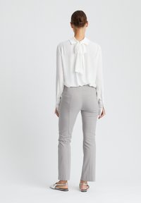 Weiße Bluse mit langen Ärmeln und Schleifendetail hinten, kombiniert mit hellgrauen, eng anliegenden Hosen und dekorativen Schuhen. Glattes Material, maßgeschneiderte Passform.